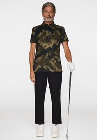 Pikétröja i kamouflage-mönster, nyanser av grönt och svart, korta ärmar, med krage. Kombinerad med svarta byxor och vita sneakers, håller en golfklubba.