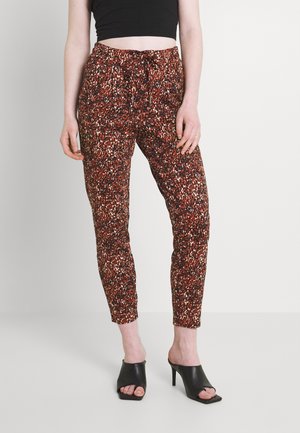 Femme portant un pantalon taille haute fuselé avec un imprimé abstrait rouge et noir et des mules à talons ouvertes noires, debout sur un fond blanc.