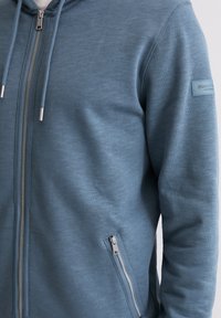 Kapuzensweatjacke mit Reißverschluss aus gedämpftem blauem Stoff, mit glatter Textur, silbernen Reißverschlüssen und einem Logo-Patch am linken Ärmel.