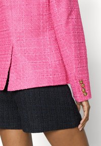 Blazer rosa con texture e bottoni dorati sui polsini, abbinato a pantaloncini neri intrecciati. Il blazer presenta un taglio dritto e una spacco laterale.
