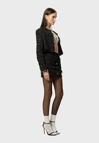 Zwarte tweed cropped jack met rafelige randen, gecombineerd met een zwarte minirok met knoopaccenten. Het model draagt witte sokken en hakken.