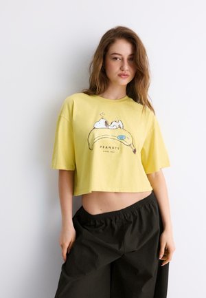 Mladá žena má na sebe žlté krátke tričko so Snoopym na banánovom motíve a voľné čierne nohavice, stojí pri bielej stene.