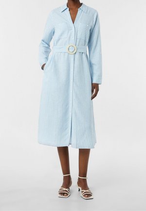 Mujer con vestido camisa midi a rayas azul claro y blanco, mangas largas, botones en la parte delantera, cinturón ancho y sandalias blancas con tiras.