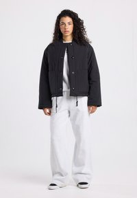 Vrouw met krullend haar in een zwarte knoopjasje, witte sweatshirt, brede witte broek en zwart-witte sneakers, staande tegen een witte achtergrond.