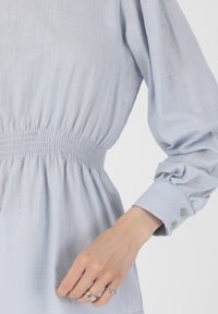 Vaaleansininen pusero, jossa on tekstuurinen kangas, rypytetty vyötärö ja puffihelmukset. Varustettu nappikiinnityksellä hihoissa ja rento leikkaus.