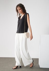 Czarny top bez rękawów z dekoltem w serek, białe spodnie o szerokich nogawkach oraz kremowa torebka na ramię w kształcie serca. Całość dopełniają czarne sandały na paski.