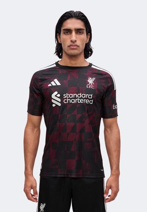 Hombre con cabello oscuro que lleva puesto un jersey de fútbol del Liverpool en negro y rojo oscuro con el logo de Standard Chartered, las rayas de Adidas y el escudo del Liverpool FC.