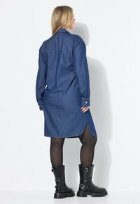 Denim-mekko, jossa on kaulus, pitkät hihat ja kaareva helma. Tummansininen väri, sileä pinta ja napitettu hihansuu, yhdistettynä paksuihin mustiin saappaisiin.
