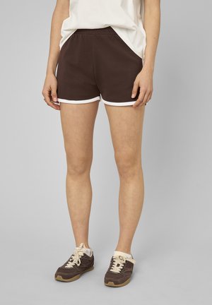 Femme portant un short de sport marron avec un liseré blanc et des baskets marron avec des lacets blancs, debout sur un fond gris.