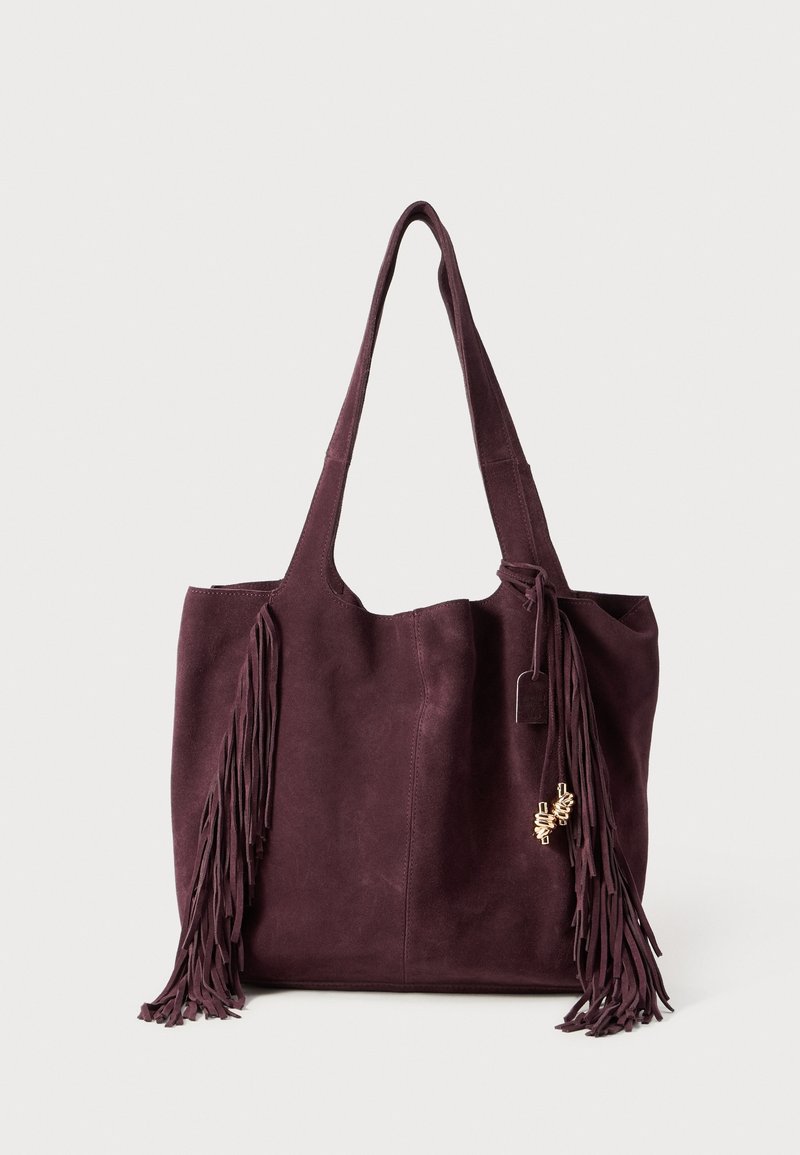 Sac à épaule en daim violet foncé avec longues anses et franges de chaque côté sur un fond clair uni.