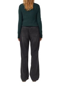 Frau mit langen Haaren, die einen dunkelgrünen Pullover und lockere schwarze Jeans trägt, steht mit dem Rücken zu einer weißen Wand.