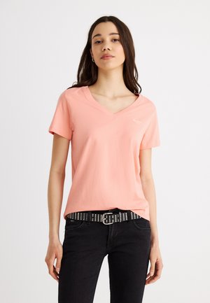 Young woman with long dark hair wears a light pink V-neck T-shirt, black studded belt, and black jeans, standing against a plain background.