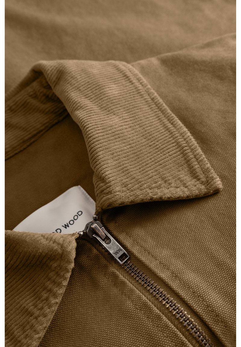 Col de veste en velours côtelé marron avec une fermeture éclair argentée. Le tissu présente des stries verticales, offrant une finition texturée. Étiquette visible à l'intérieur du col.