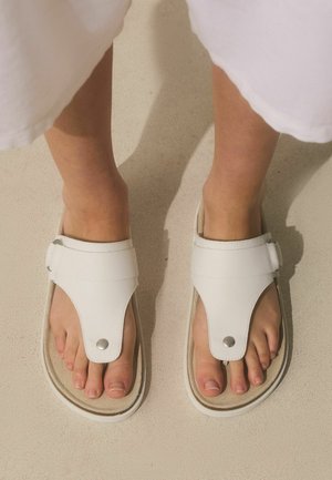 Nohy v bielych sandáloch s otvorenou špičkou, širokými remienkami a kovovými gombíkmi na svetlej podlahe.