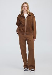 Brązowa kurtka zapinana na zamek z miękką, zamszową fakturą oraz pasującymi brązowymi spodniami o szerokich nogawkach. Posiada białą wyściółkę kołnierza oraz haftowane logo.