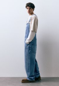 Lichtblauwe denim tuinbroek met verstelbare schouderbanden, zakken en een losse pasvorm, gecombineerd met een beige shirt met lange mouwen en bruine schoenen.