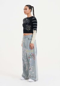 Maglietta nera a maniche lunghe e corta con strisce bianche, con grafica di un teschio; jeans baggy blu chiaro con ricami floreali colorati. Calzature bianche.