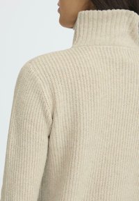 Beige ribbestrikket genser med høy krage, med et strukturert strikkemønster og myk overflate, vist fra baksiden.