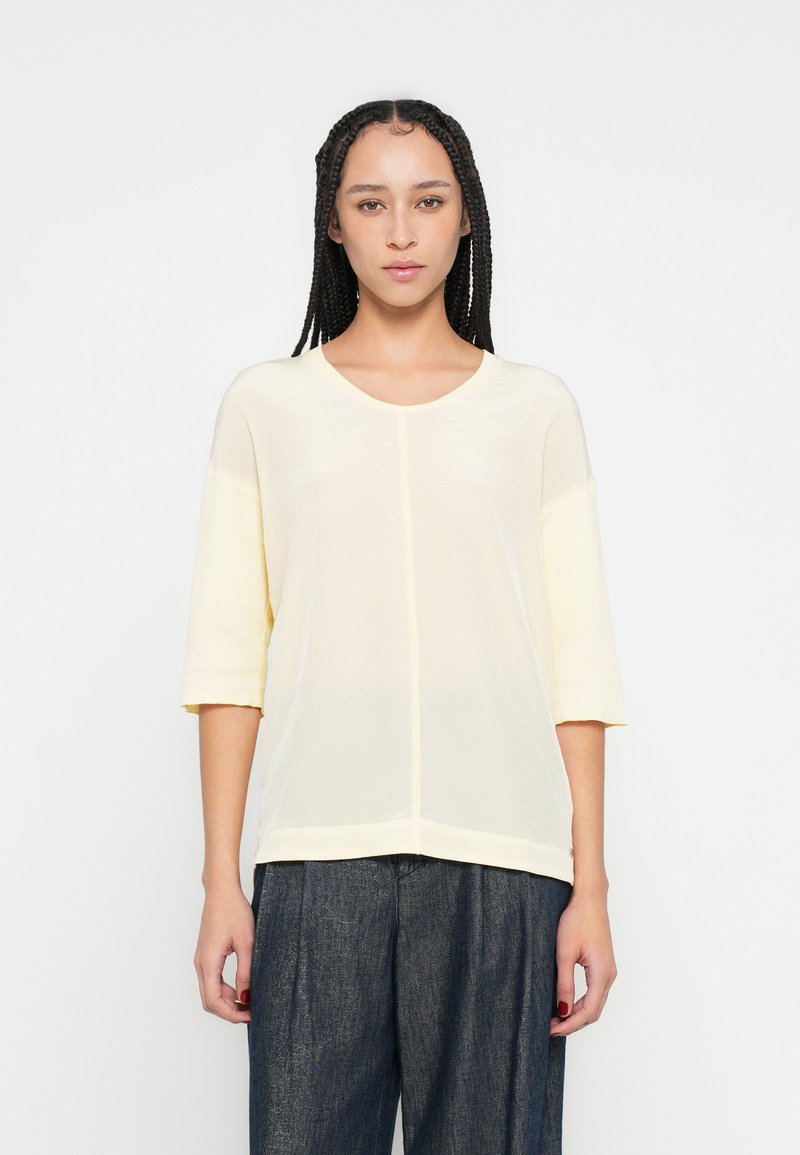 Top galben deschis, larg, cu mâneci trei sferturi și decolteu rotund. Țesătură netedă cu o cusătură ce se întinde vertical în mijloc.