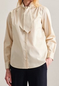 Cream-colored blouse with a tie collar, long sleeves, and button front. Made from smooth, lightweight fabric with subtle sheen.