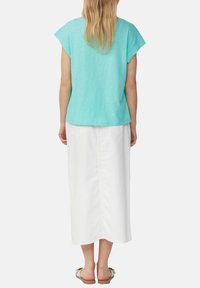 Vrouw met lang blond haar, gekleed in een turquoise top met korte mouwen, een witte enkellange rok en witte sandalen, staand met haar rug naar de kijker.
