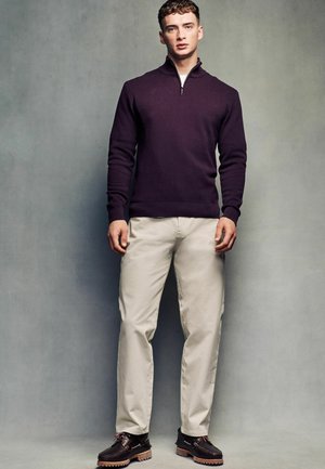 Violetti vetoketjuhuppari, jossa on teksturoitu kuvio, beige housut ja tummanruskeat kengät, joissa on vaaleanpunaiset nauhat ja kestävät pohjat, seisoo harmaata taustaa vasten.