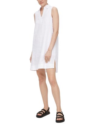 Witte mouwloze linnen jurk met een kraag, zijslitsen en een relaxte pasvorm. Gepaard met zwarte sandalen met banden en minimale accessoires.