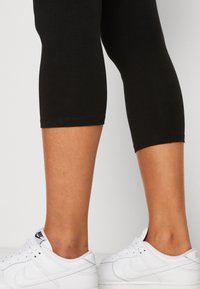 Fekete capri leggings, puhából készült, fehér sportcipőkkel párosítva, amelyeken texturált felület és egyedi logó található az oldalon.