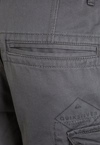 Grå tyg cargo shorts med diagonala sidofickor, horisontell dragkedjeficka och ett märkespatch på sidan som visar logodetaljer.