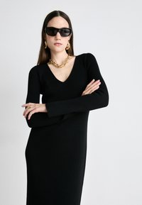 Black ribbed knit dress with a V-neck and long sleeves. Accented with gold chain necklace and bold sunglasses.
