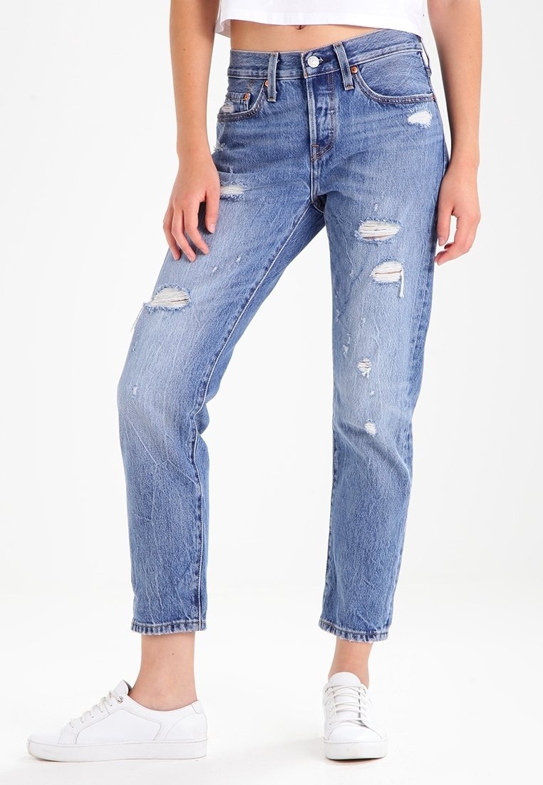 Jean en denim bleu clair avec des déchirures usées et des ourlets effilochés, doté d'une taille haute et d'une coupe droite. Associé à des baskets blanches.