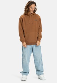 Brown cotton hoodie with drawstring, front pocket, and logo patch; paired with loose light blue jeans and white sneakers.