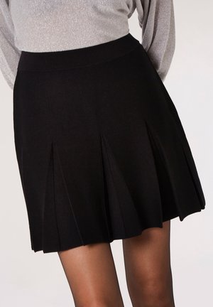 Mini-jupe noire plissée portée avec un haut argenté à manches longues, montrant le bas du torse et le haut des jambes sur un fond uni.