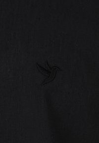 Sort stof har en tekstureret overflade med et broderet kolibri-logo placeret på venstre side.