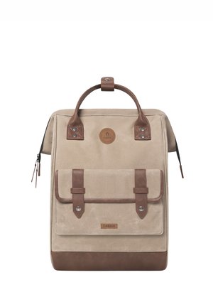 Beige canvas backpack with brown leather accents, dual handles, front pocket with flap, and zipper closure. Circular logo and rivets visible.