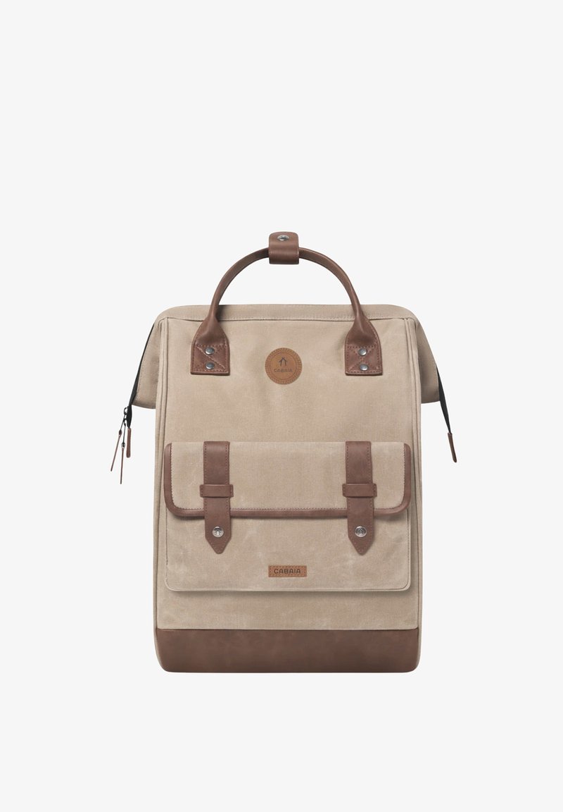 Beige canvas backpack with brown leather accents, dual handles, front pocket with flap, and zipper closure. Circular logo and rivets visible.