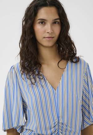 Žena s dlhými kučeravými tmavými vlasmi, oblečená v modrej, bielej a čiernej vertikálne pruhovanej blúzke s výstrihom do V a nariaseným predným detailom.