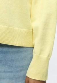 Personne portant un pull jaune pâle avec poignets et ourlet côtelés, et un jean en denim bleu clair, le bras détendu le long du corps.