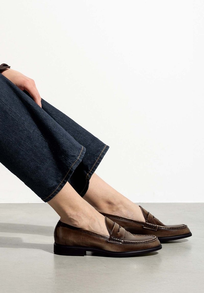 Mocassins en cuir marron avec un design cousu et des détails à l'avant, associés à un pantalon en denim foncé, sur un fond de sol neutre.