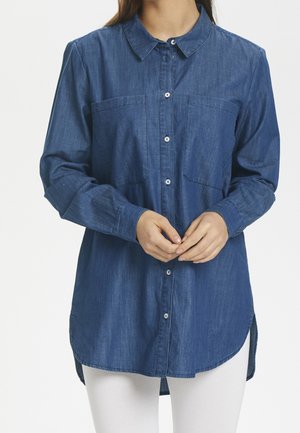 Woman wearing long-sleeve blue denim shirt with front pockets and white buttons, paired with white pants, hands loosely clasped at waist.