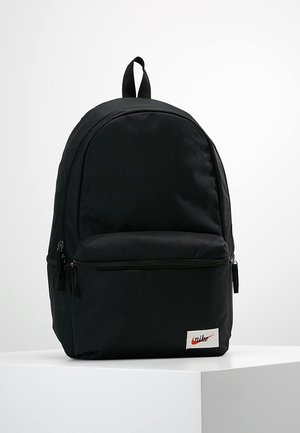 Czarny plecak Nike z przednią kieszenią na suwak, górnym uchwytem i naszywką z logo na białej powierzchni na jednolitym tle.