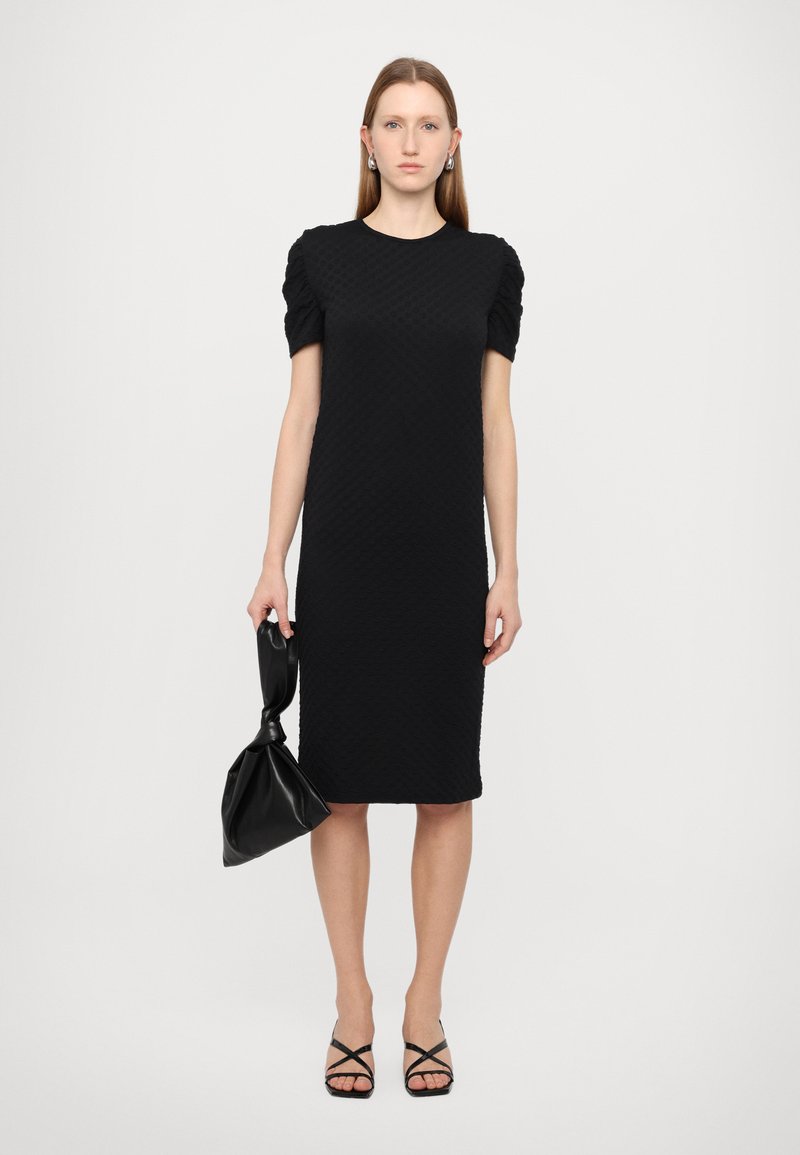 Vestido negro texturizado con mangas cortas abullonadas, hasta la rodilla. Combinado con un bolso negro anudado y sandalias de tacón alto negras con tiras.