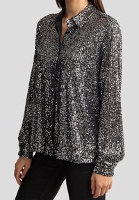 Chemise noire à sequins avec col, manches longues, patte de boutonnage, et une texture brillante qui crée un effet scintillant.