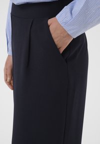 Main dans la poche d'une jupe bleu marine foncé portée avec une chemise à manches longues à carreaux bleu clair sur un fond uni.
