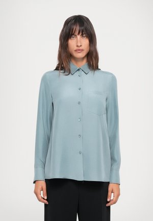 Light teal long-sleeve shirt, button-up design with a pointed collar and a chest pocket. Smooth texture, paired with black trousers.