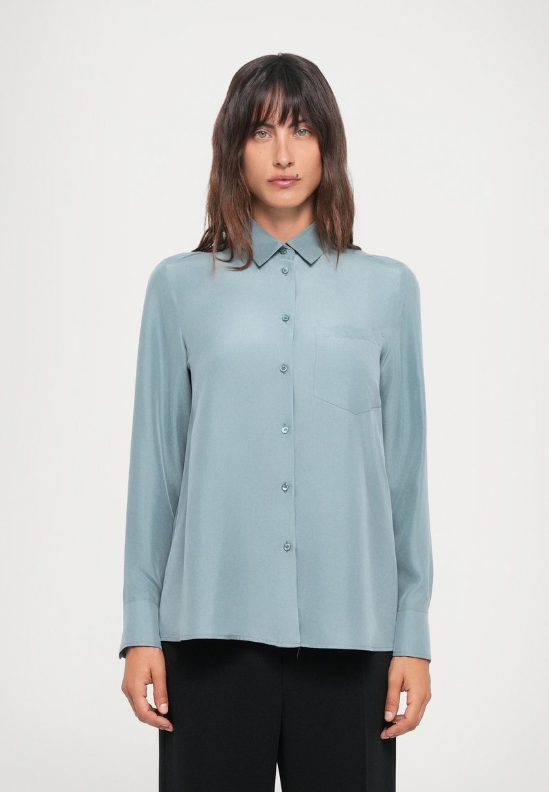 Light teal long-sleeve shirt, button-up design with a pointed collar and a chest pocket. Smooth texture, paired with black trousers.