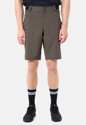 Homme portant un short marron jusqu'aux genoux, des chaussettes noires à rayures blanches et des baskets noires, debout devant un fond uni.