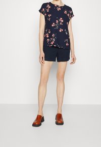 Top corto a maniche corte blu navy con motivo floreale rosa e verde, abbinato a pantaloni corti scuri e zoccoli marroni con tacco a blocco.