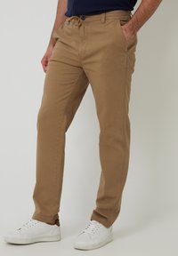 Chinos beige con un corte ajustado, hechos de algodón, que cuentan con bolsillos laterales, cierre de botón y un diseño de pierna recta. Llevados con zapatillas blancas.