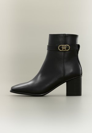 Bottine noire en cuir avec talon bloc et boucle dorée décorative sur le côté sur fond clair.